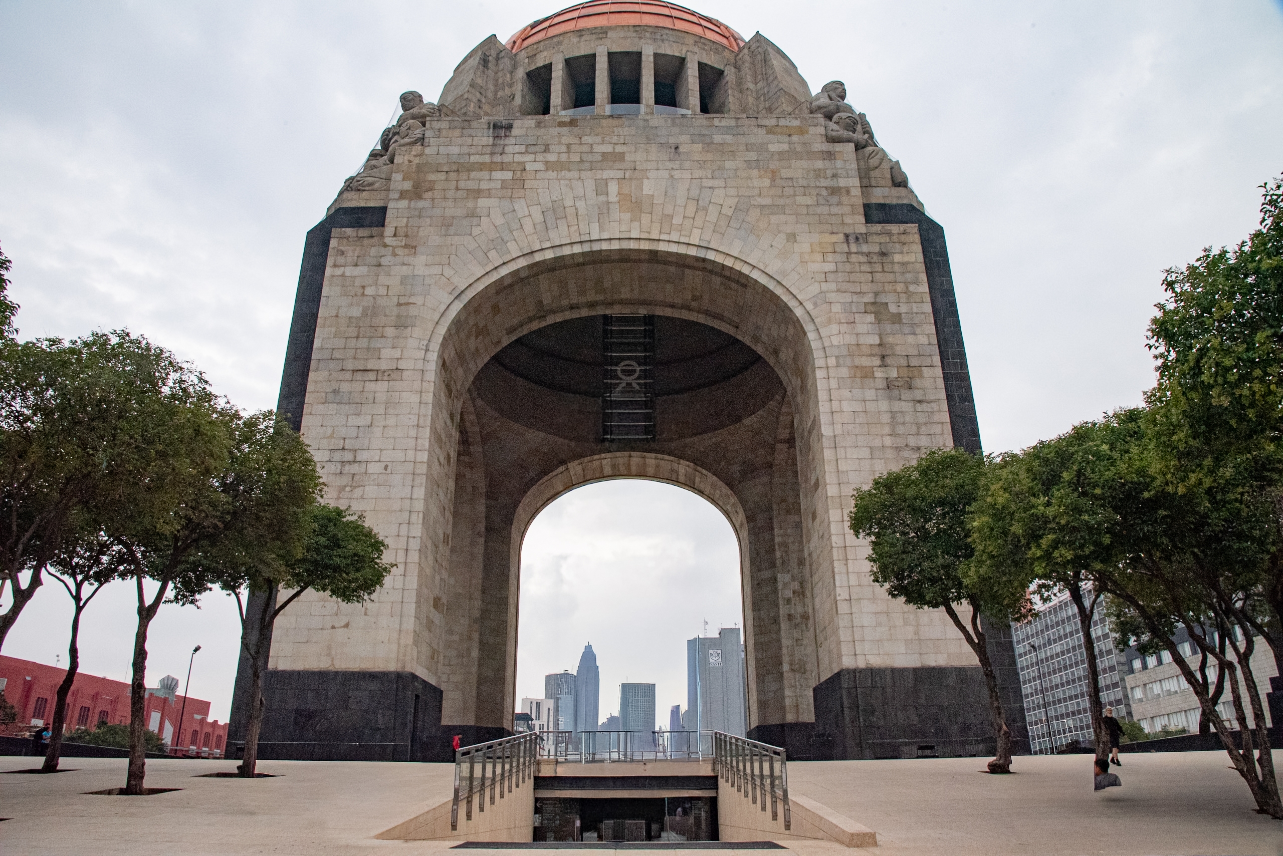 The Monument to the Revolution in Mexico City, a large, stone archway with a bronze dome, viewed from a pedestrian plaza. Modern skyscrapers are visible through the arch. This iconic landmark is a prime location for MICE industry events and incentive travel.
