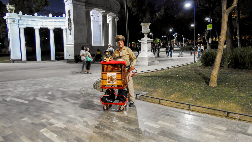 A traditional Mexican organ grinder, known as a 'cilindrero,' stands with his instrument on a street at night in Mexico City. The background shows a public park with people walking and a historic stone archway. This image captures the unique cultural experiences available in Mexico City, perfect for MICE industry event entertainment and incentive travel.