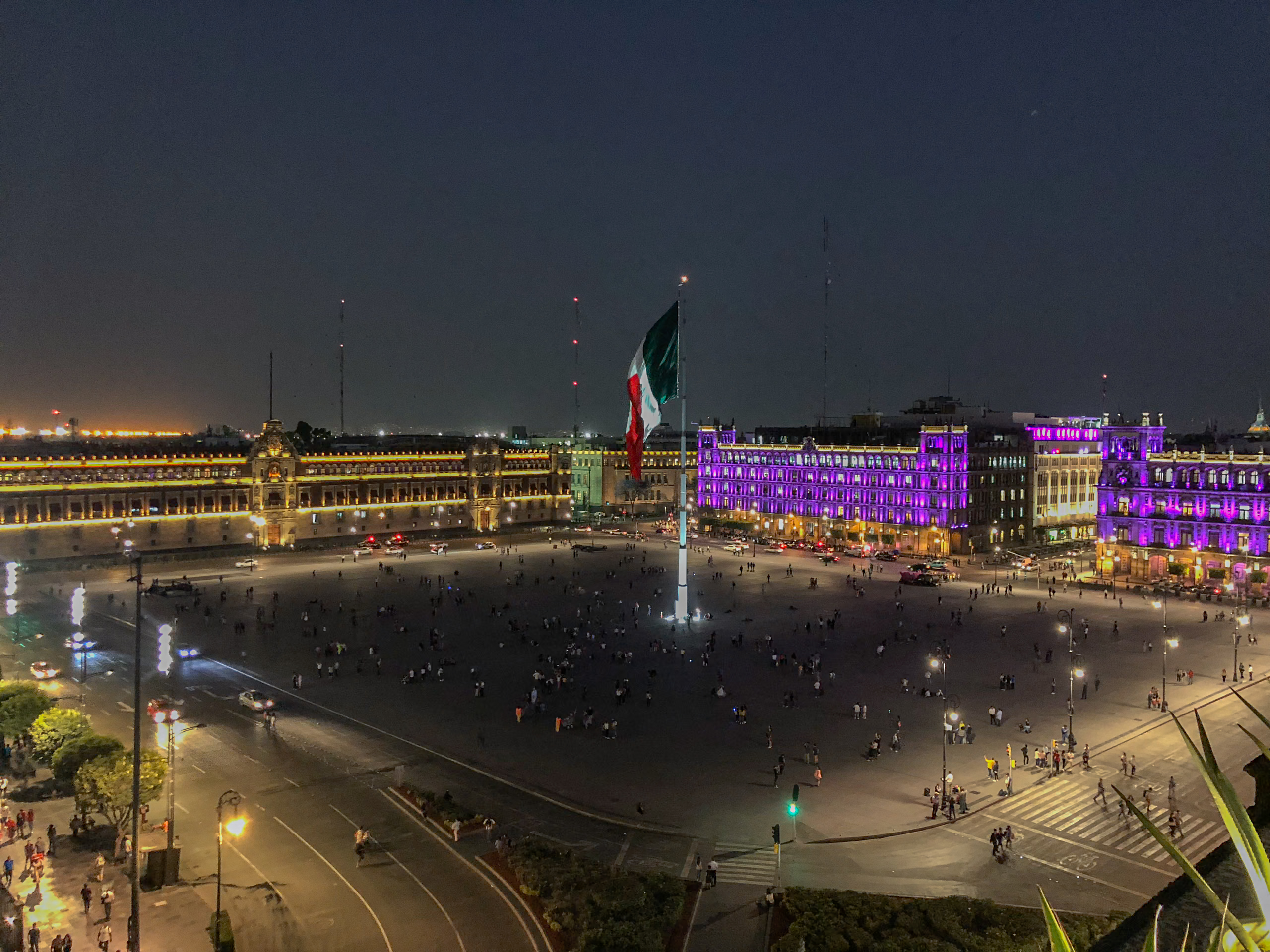The Zócalo at night in Mexico City, a massive public square illuminated by streetlights and the glowing lights of surrounding historic buildings, including the National Palace. A large Mexican flag stands in the center of the square, where people are gathered. This vibrant scene is a perfect place to visit for MICE industry events and incentive travel.