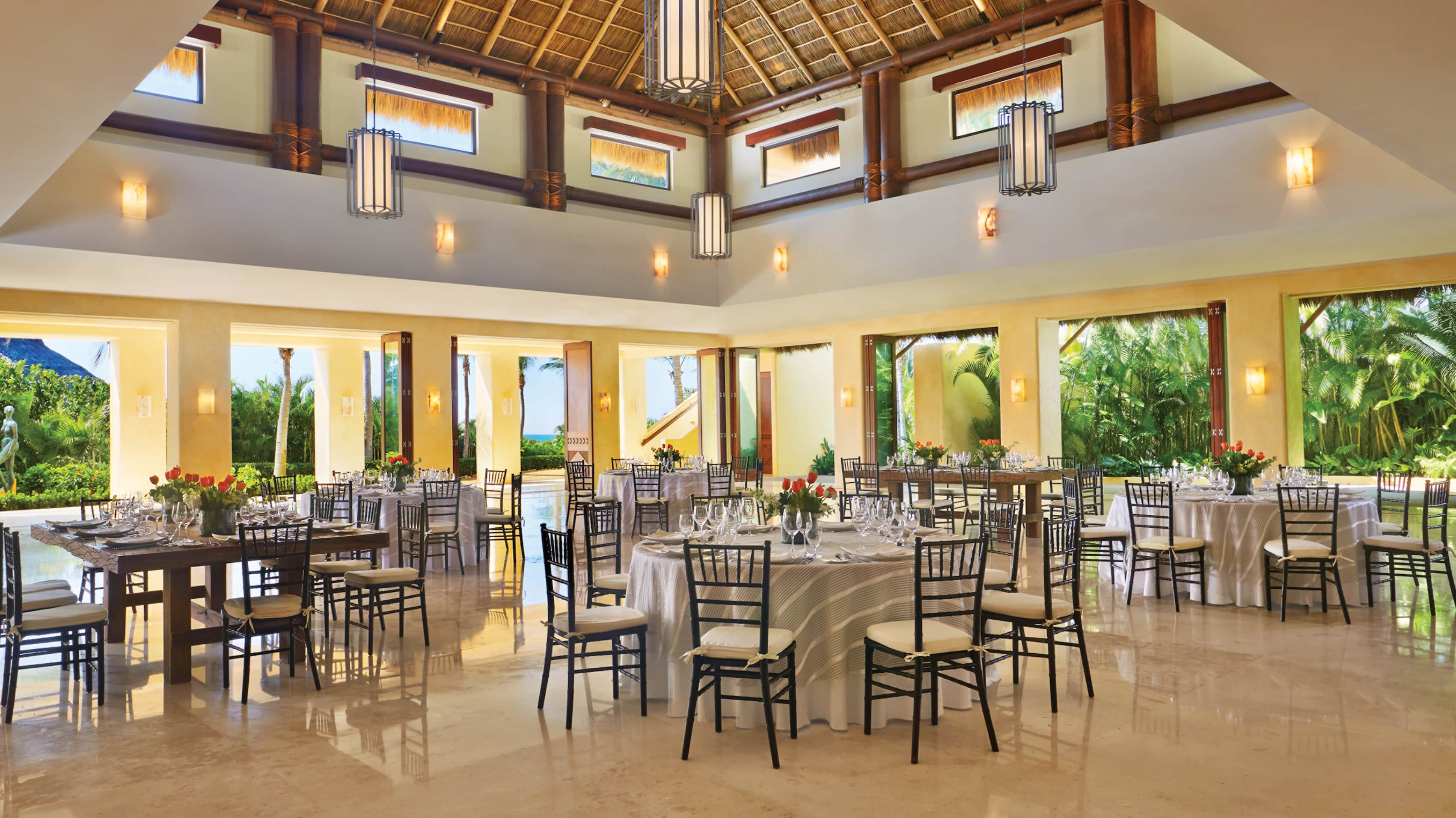 Elegant banquet hall setup at Four Seasons Resort Punta Mita, a luxury MICE venue in Mexico for meetings, incentives, conventions, and exhibitions, featuring thatched ceiling with pendant lights, round and rectangular tables with white linens, black chairs, floral centerpieces, and large windows overlooking tropical gardens and ocean views ideal for corporate events and incentive travel banquets in Riviera Nayarit.