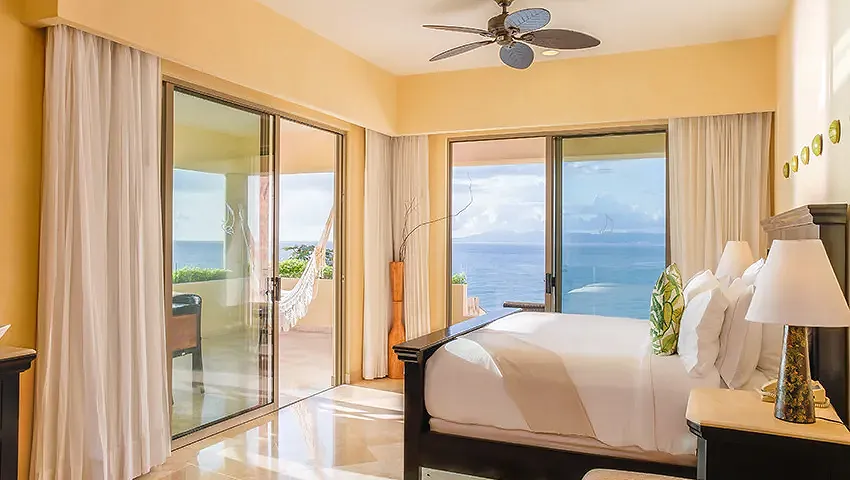 Luxurious ocean-view bedroom at Garza Blanca Preserve Resort & Spa, a premier MICE venue in Mexico for meetings, incentives, conventions, and exhibitions, featuring a king-size bed with white linens and green accents, ceiling fan, sheer curtains, bedside lamps, and balcony with hammock overlooking the Pacific Ocean and tropical landscape ideal for attendee relaxation during incentive travel programs in Puerto Vallarta.