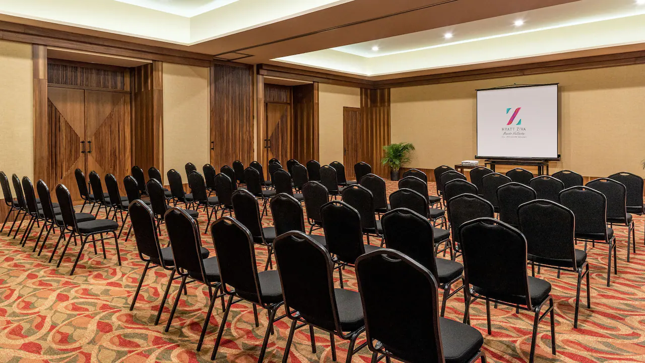 Theater-style meeting room setup at Hyatt Ziva Puerto Vallarta, a leading MICE venue in Mexico for meetings, incentives, conventions, and exhibitions, featuring rows of black padded chairs arranged in semi-circles facing a projection screen with Hyatt Ziva logo, wooden paneled walls, double doors, potted plant, and patterned carpet ideal for corporate presentations and breakout sessions in an all-inclusive resort environment.